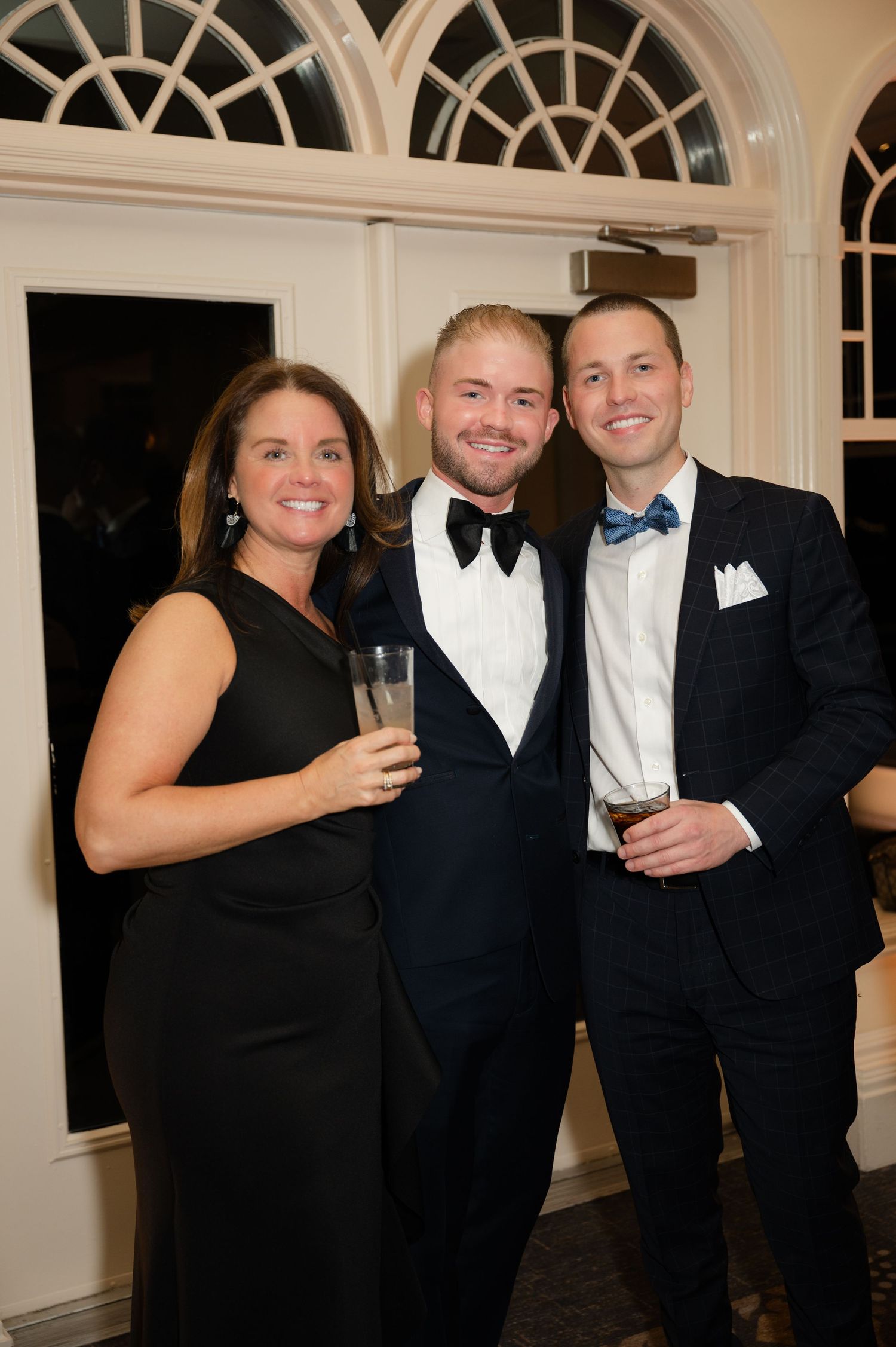 Well-dressed wedding guests in formal attire enjoying drinks at an elegant event with arched windows in the background.