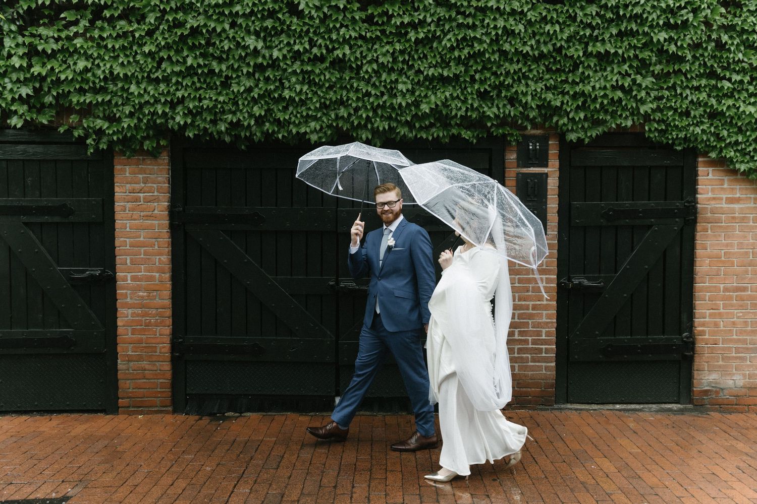 A couple shares a clear umbrella while standing on brick pavement against an ivy-covered wall on a rainy day.