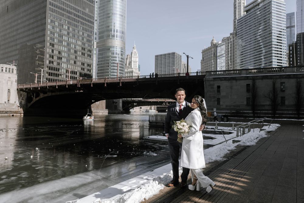 A couple stands in winter along Chicago's riverwalk with snow and ice, framed by a bridge and urban skyscrapers.