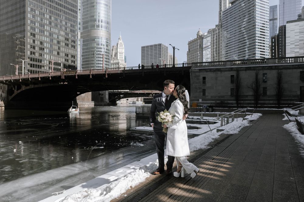 A couple in winter attire embraces along the snowy Chicago Riverwalk with skyscrapers and bridge in the background.