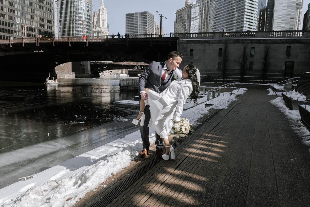 A couple shares a romantic moment on a winter riverside path in Chicago with snow and urban buildings in the background.