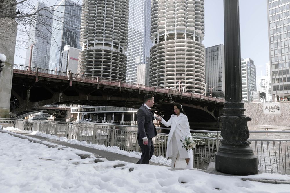 Two figures stand in winter snow along Chicago Riverwalk with Marina City towers and downtown skyscrapers in background.