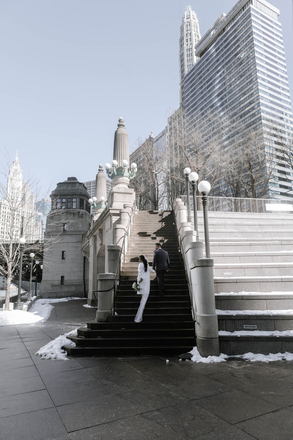Snow-dusted stone staircase with decorative columns leads up to modern skyscrapers in an urban setting.