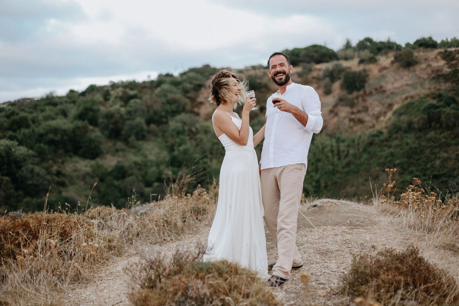 A couple in white outfits poses together on a hillside path during a sunset photoshoot in dry grassland terrain.