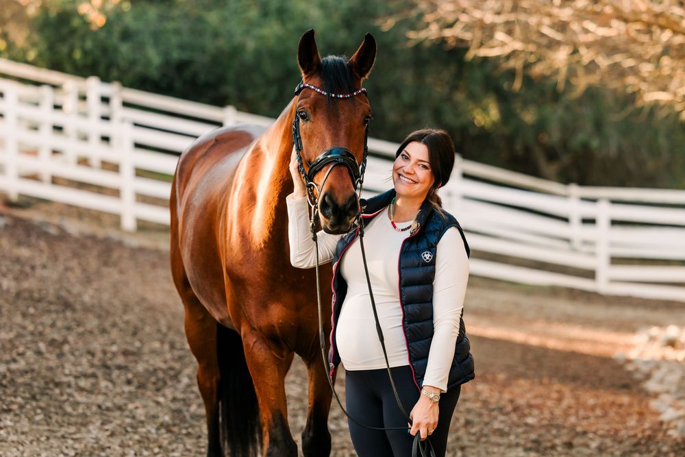 A beautiful bay horse stands beside someone on a farm with white fencing visible in the background.