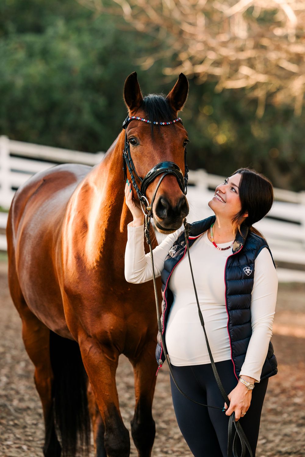 A tender moment between a rider in a puffy vest and their bay horse against a warm sunset backdrop.