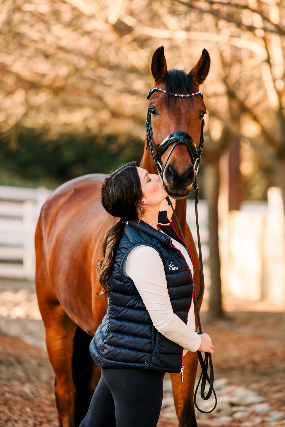 A person in a black puffy vest stands with a beautiful bay horse in a fall setting with golden leaves.