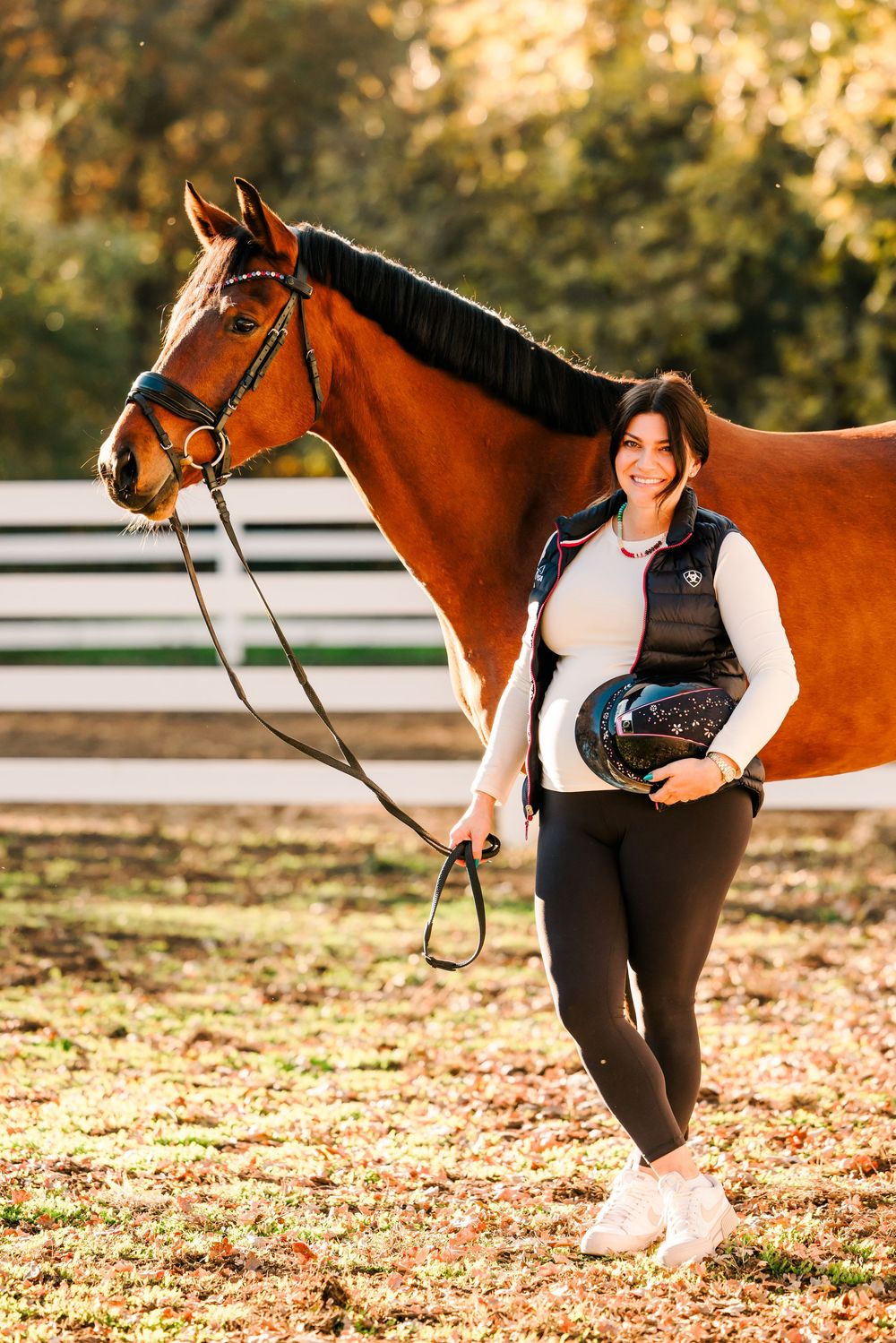 Beautiful bay horse and equestrian posing together in autumn golden light with white fencing in background.