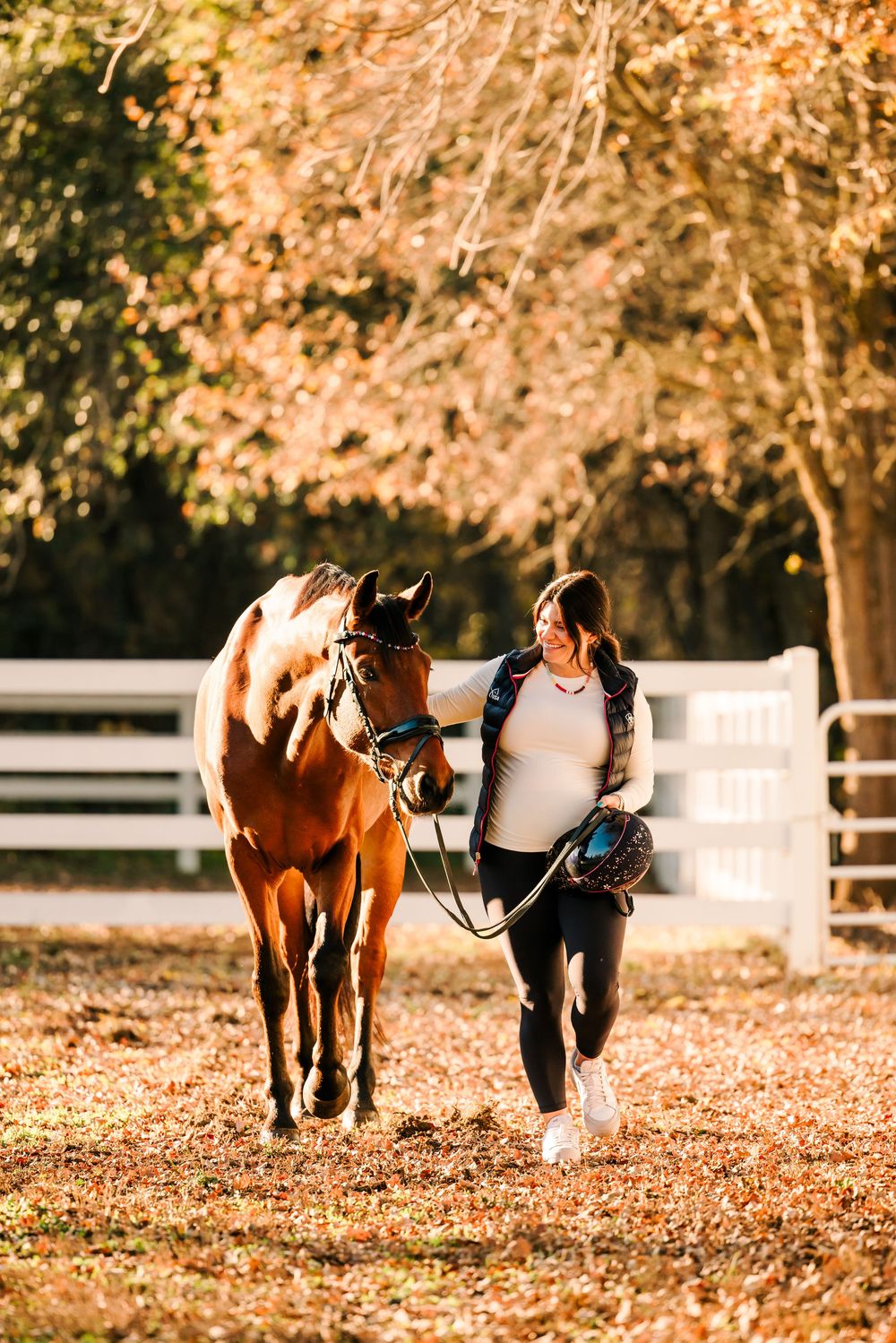 A person leads a chestnut horse through autumn leaves on a path bordered by white fences at sunset.