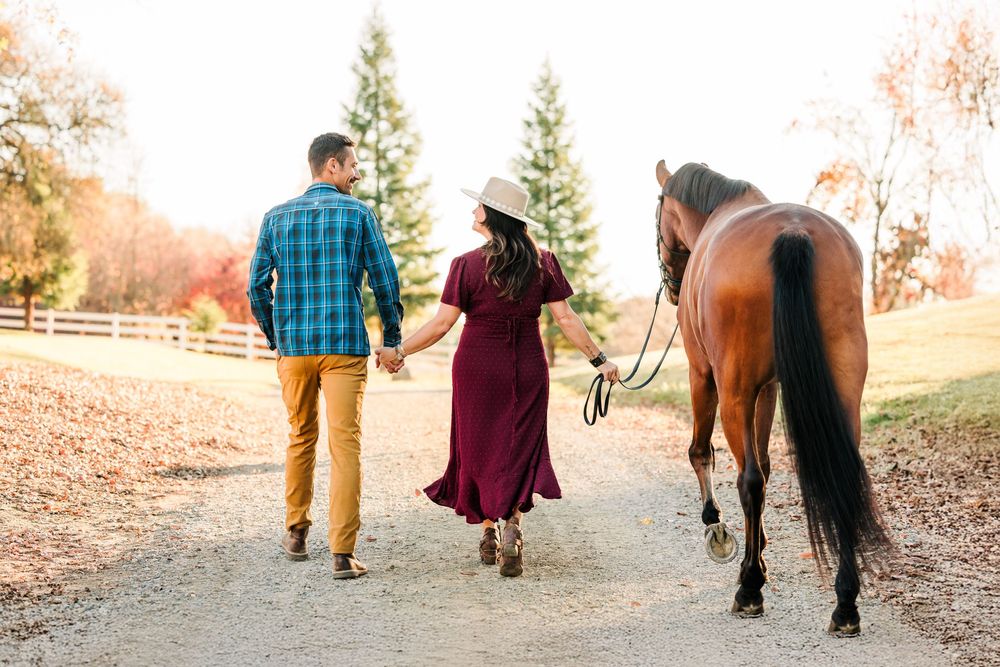 A couple walks hand in hand on an autumn path with their horse following behind them.