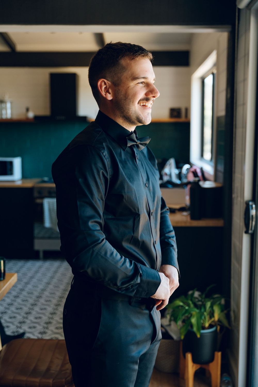 Groom in a classic black suit and bow tie smiles while looking out a window in a modern interior setting.