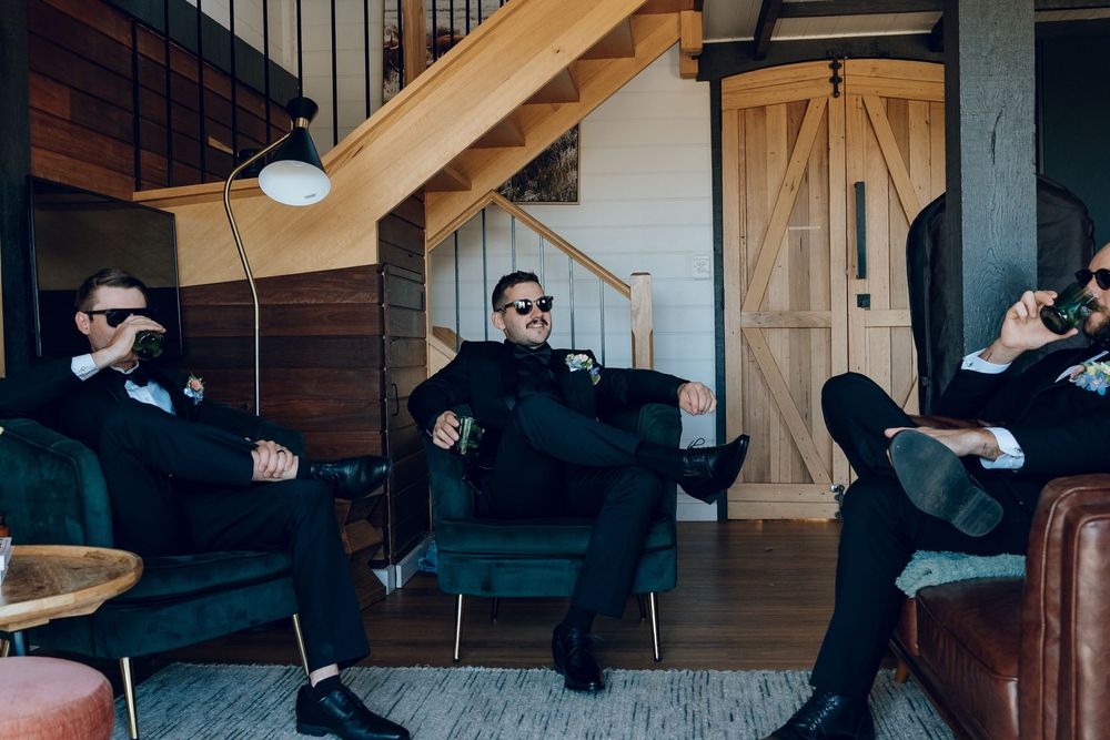 Wedding party relaxes on dark leather couches in a rustic wooden interior.