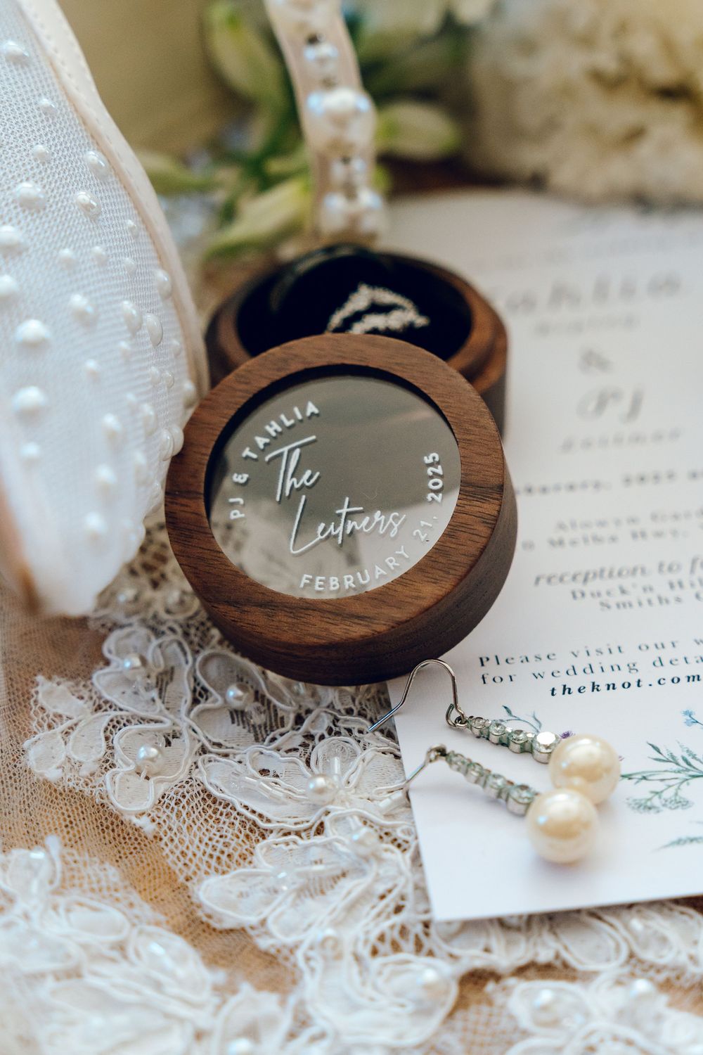 Wooden ring box with custom engraving sits on delicate lace with pearls and wedding details.