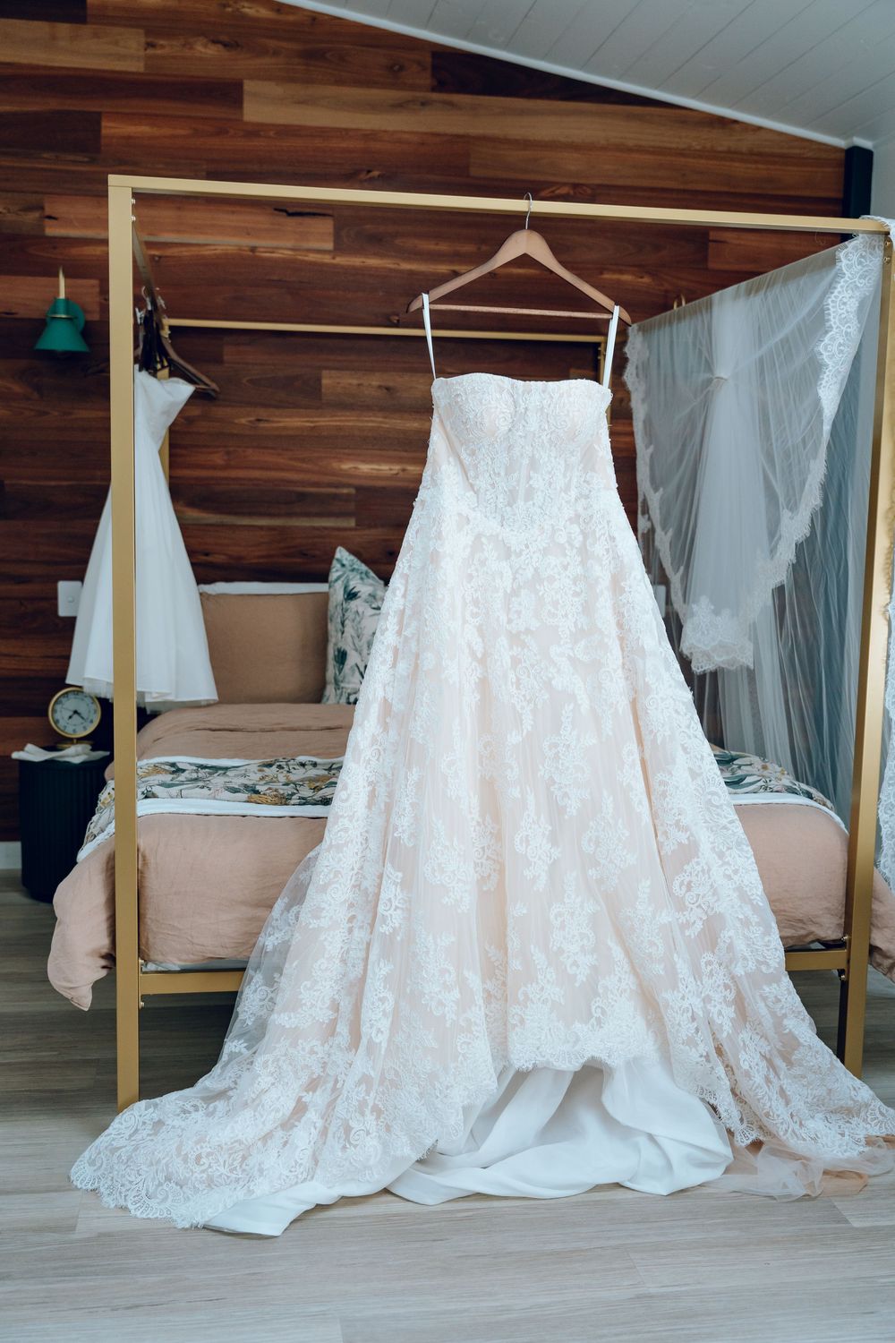 Elegant white lace wedding dress hangs on display against wooden wall in bridal suite.