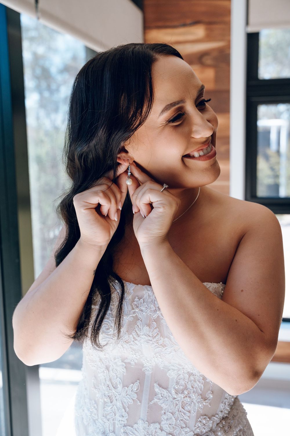 Bride putting on earrings while wearing lace detailed wedding dress.