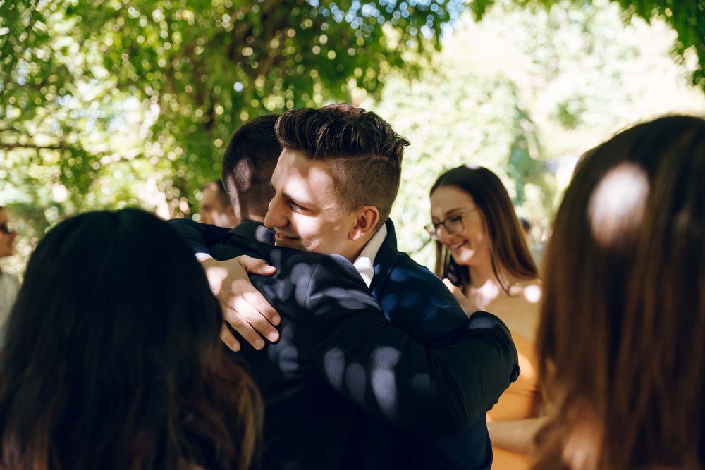 Wedding guests enjoy a casual outdoor gathering under a canopy of green trees.