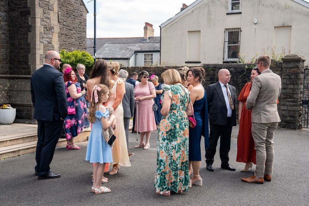 Guests in colorful formal attire gather outside a stone building during a social celebration.