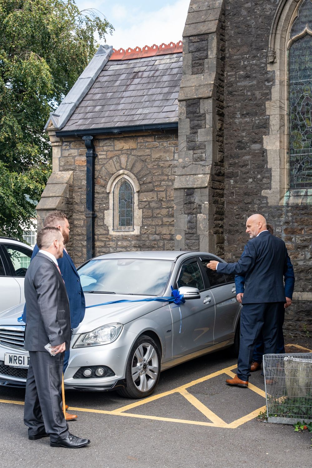 Two men in suits standing next to a silver Mercedes-Benz car parked outside a stone church building.