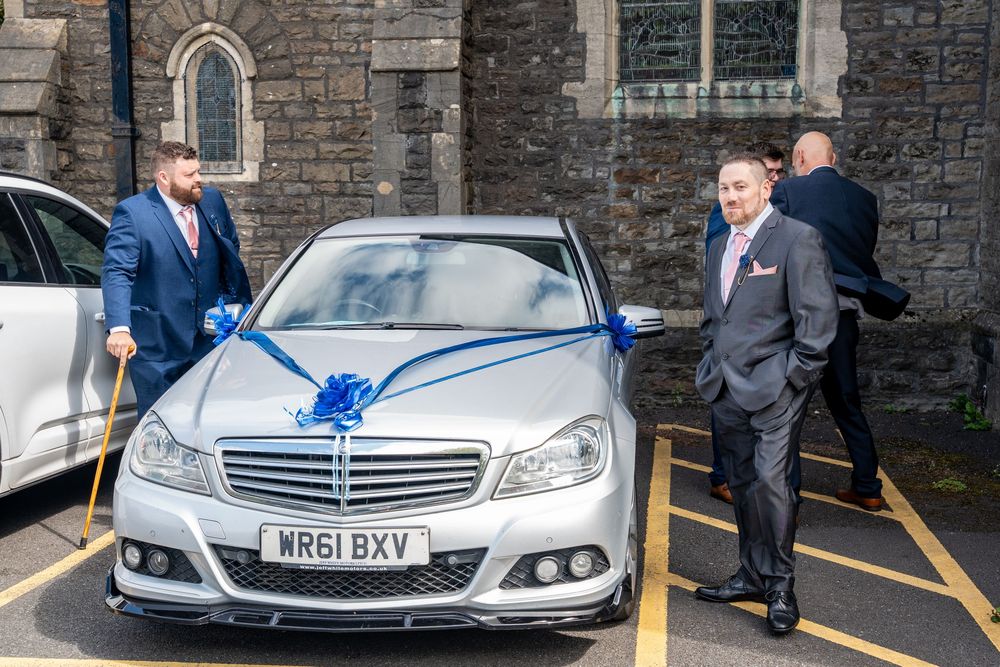 A silver Mercedes-Benz decorated with a blue bow sits parked outside a stone church building.