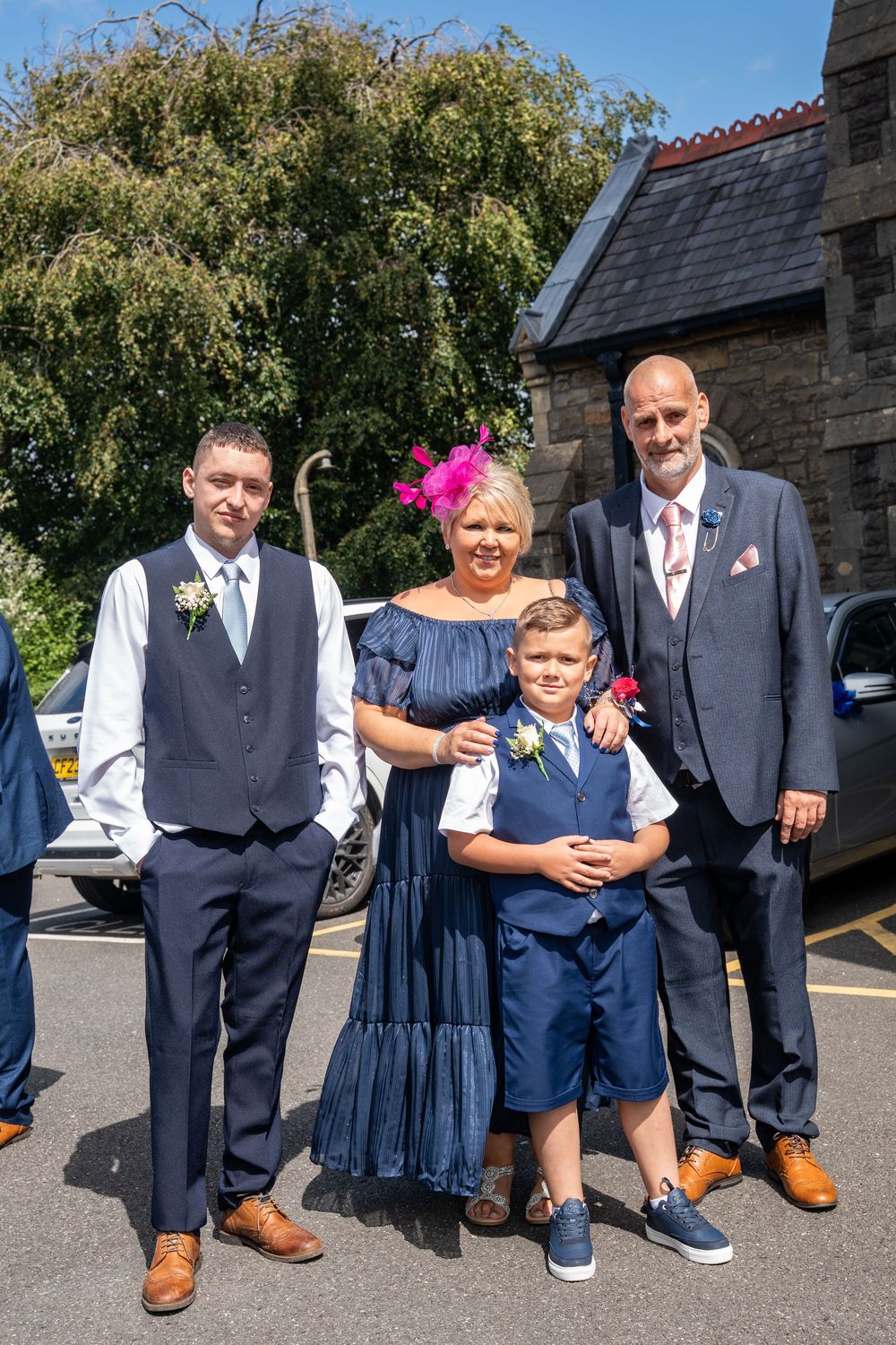 A group of four people in formal attire pose together outdoors near a stone building on a sunny day.