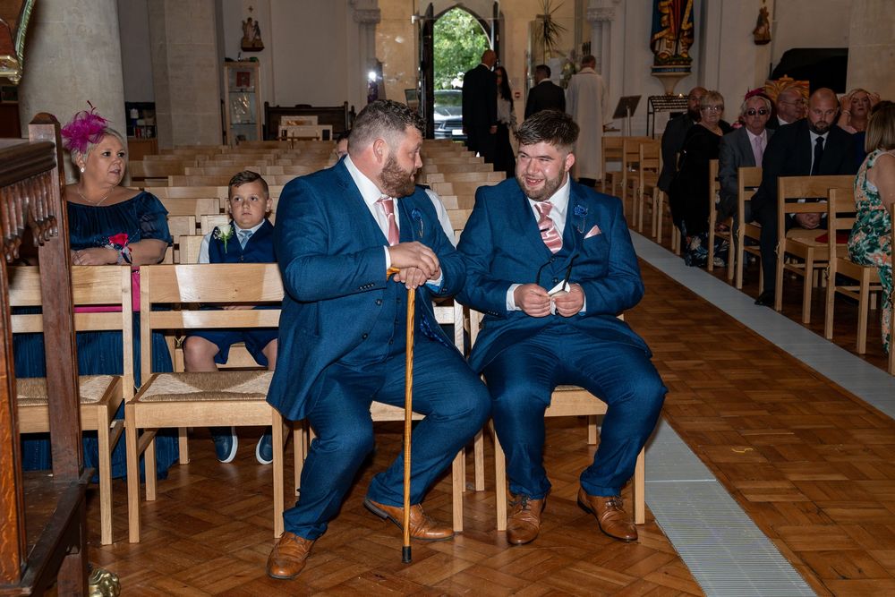 Two people in matching blue suits sit in wooden church pews during a ceremony, one holding a walking stick.