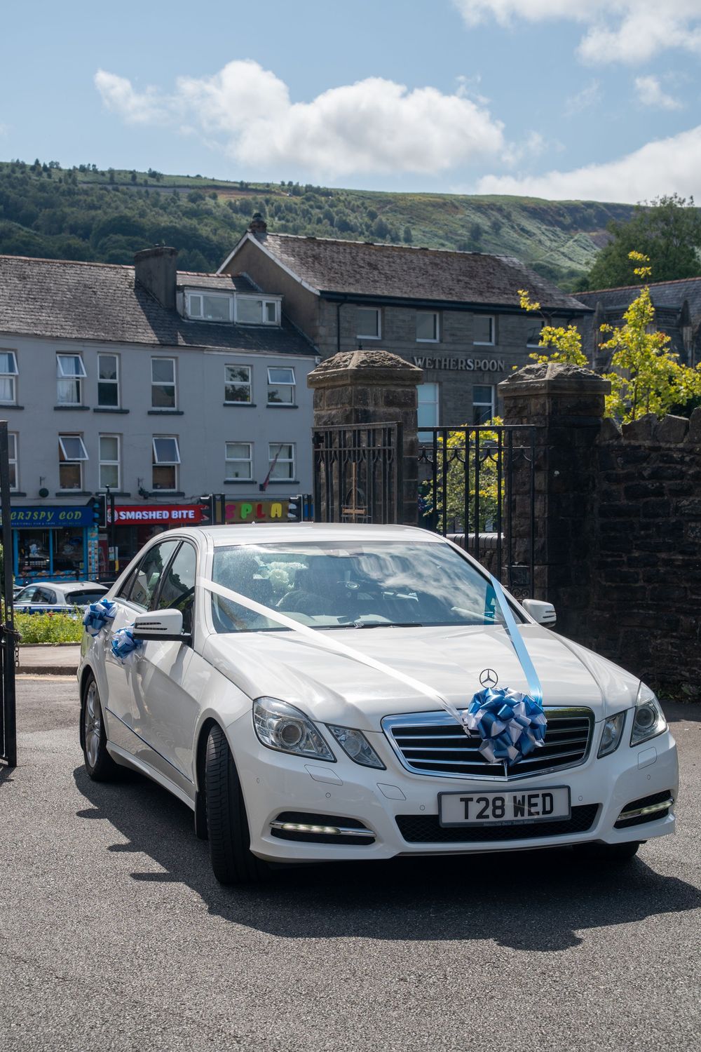 A white Mercedes-Benz E-Class decorated with blue ribbons for a wedding sits parked in front of buildings on a sunny day.