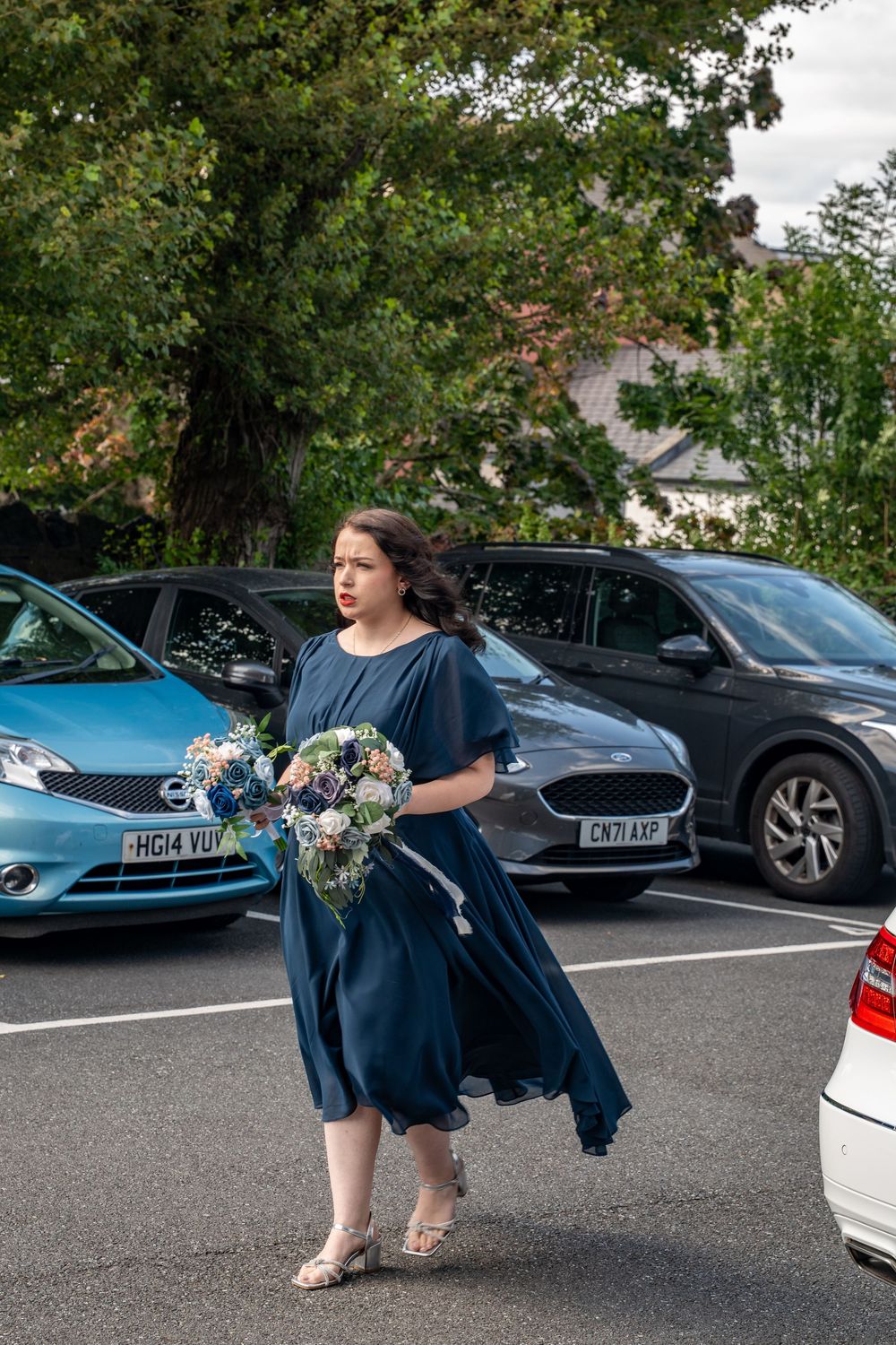 A person in a navy blue dress carries a floral bouquet through a parking lot with cars on a cloudy day.
