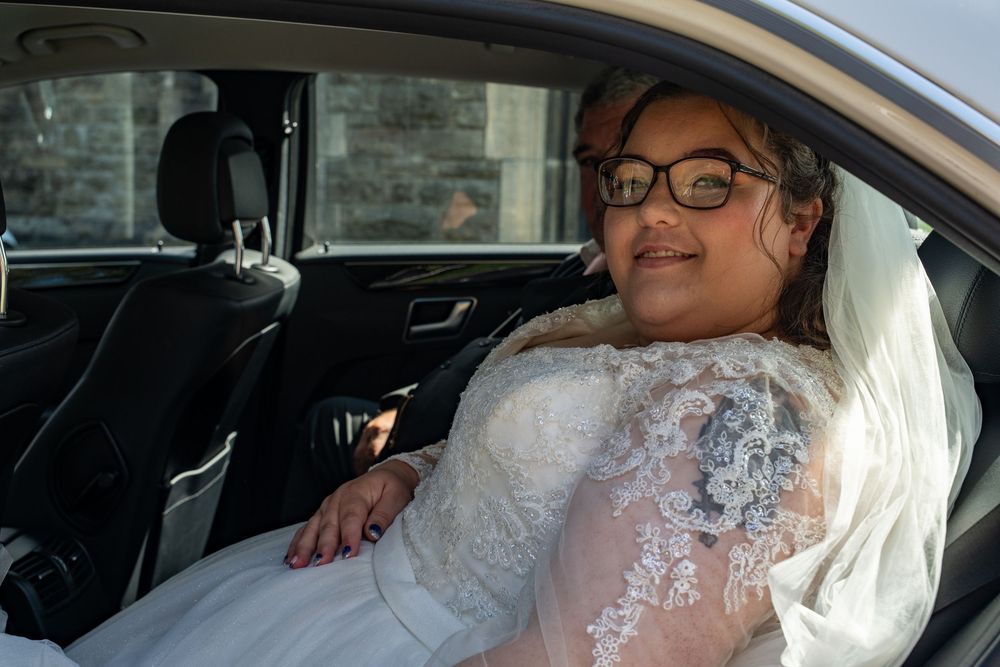 A bride wearing a lace wedding dress sits in the passenger seat of a car during her wedding day.