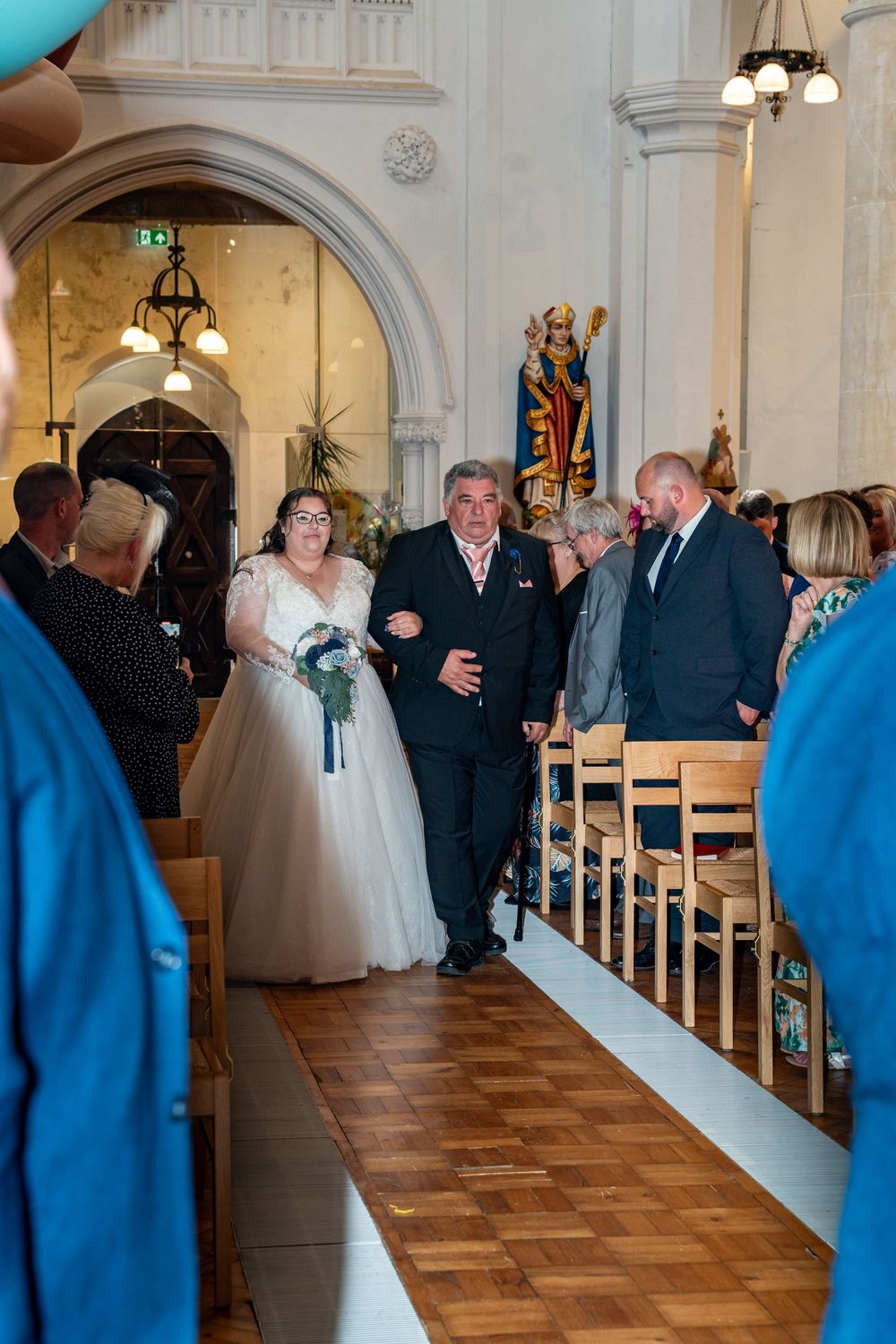 A wedding ceremony takes place inside a beautiful church with arched doorways and wooden floors as guests look on.