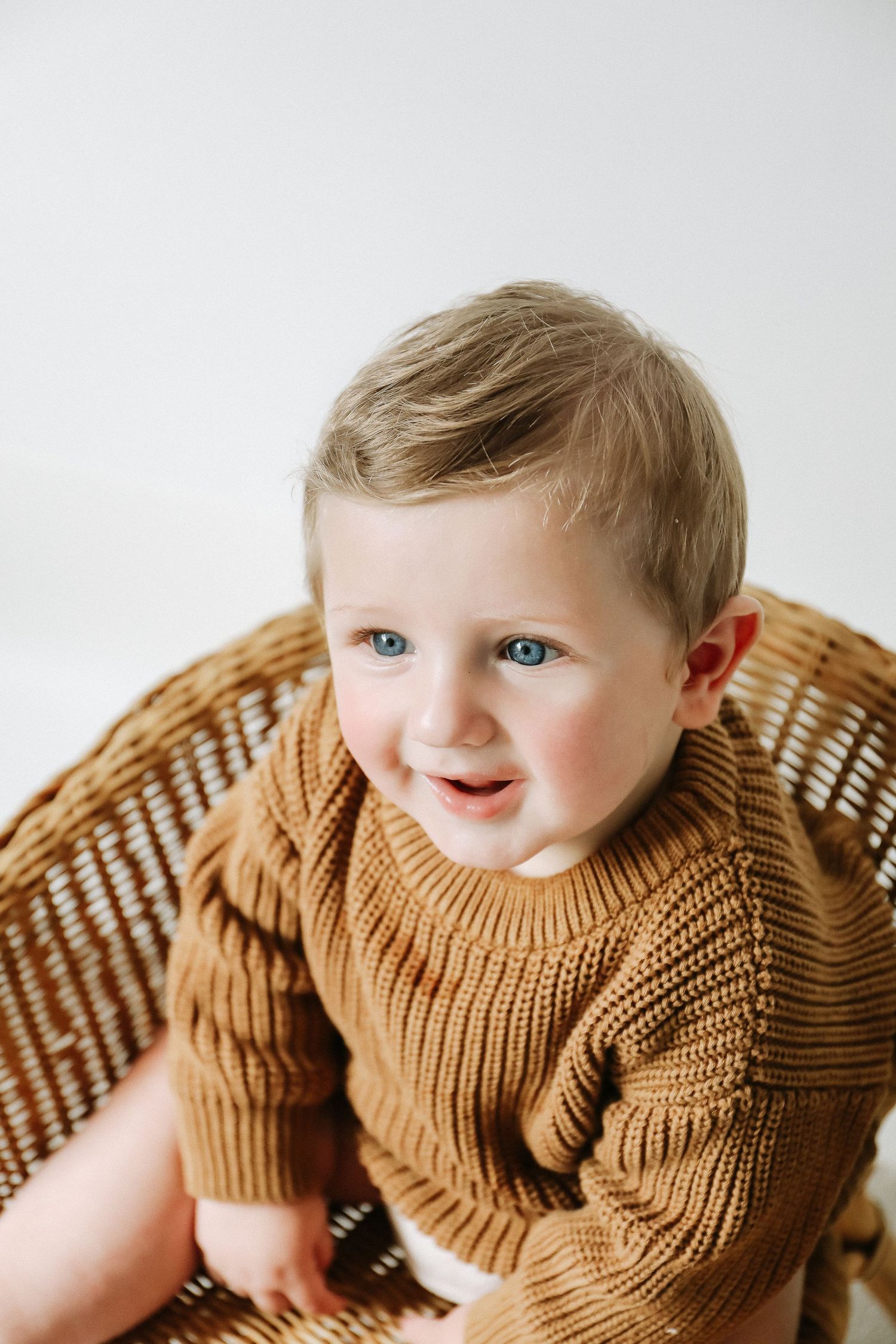 Show a baby wearing a brown knit sweater sitting in a wicker chair against a white background during birthday shoot in Nottingham