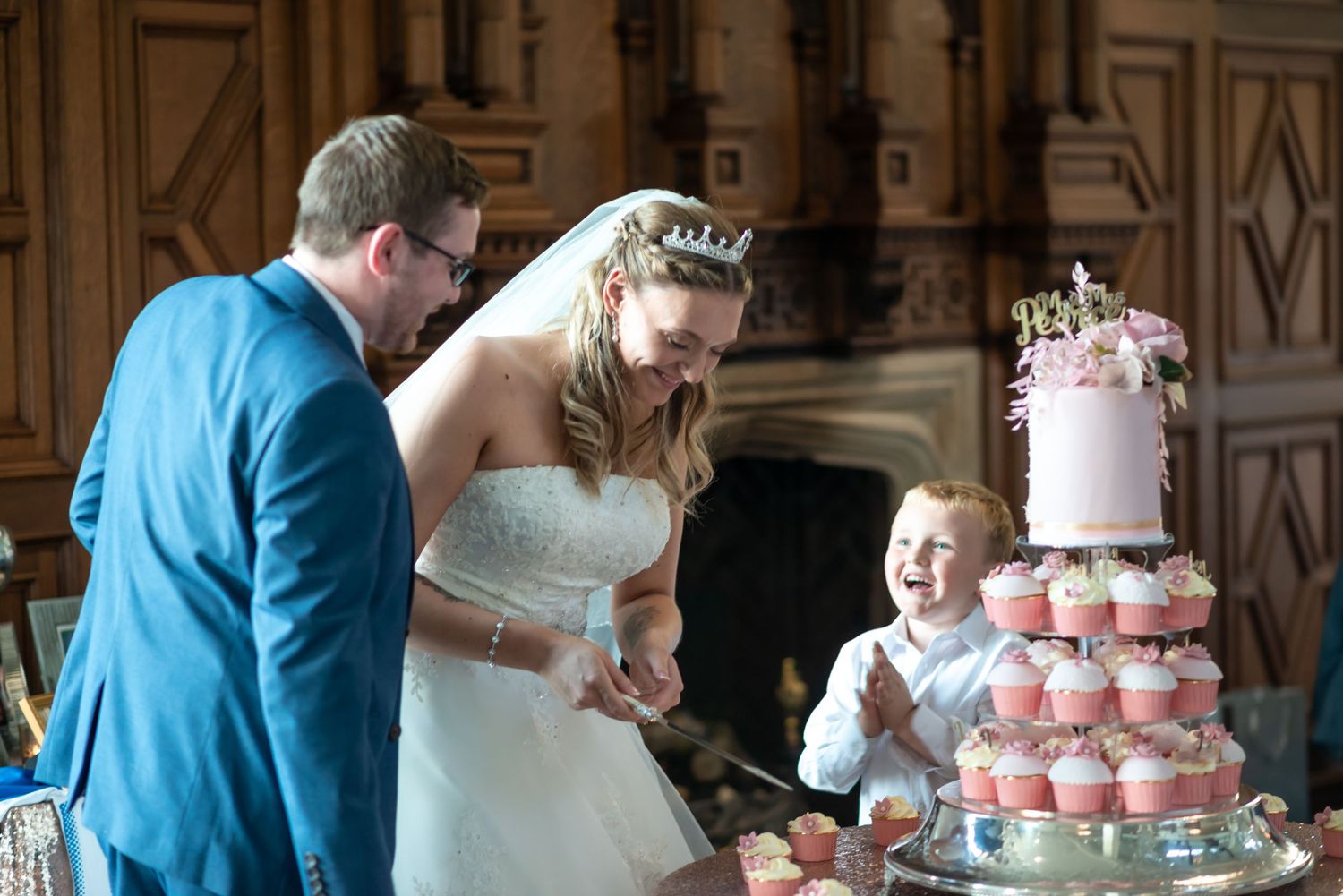Wedding couple cutting into a pink tiered cake with cupcakes while a young guest watches with excitement.