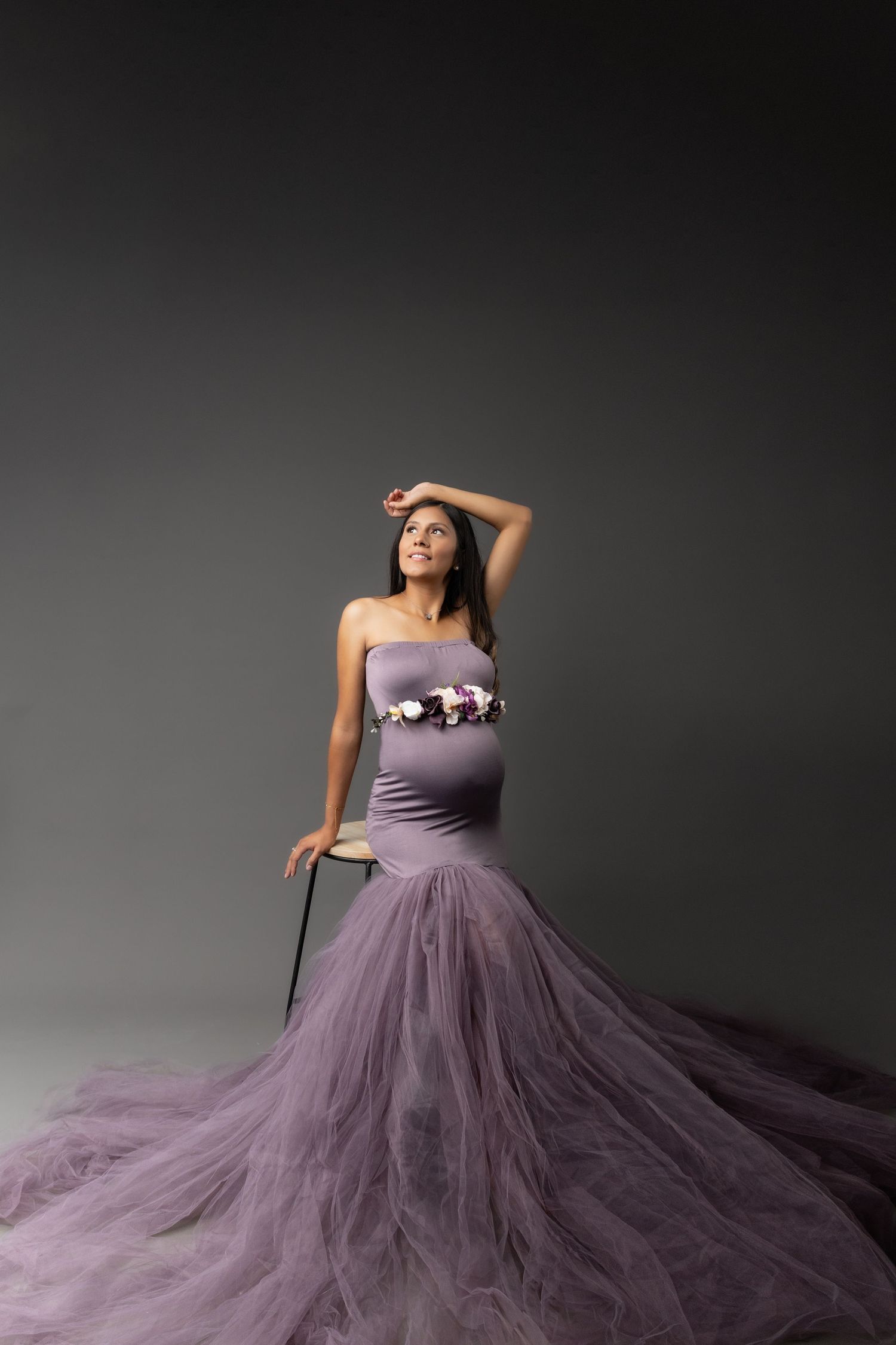 A person poses elegantly in a flowing purple tulle gown with a fitted bodice against a dark gray studio backdrop.
