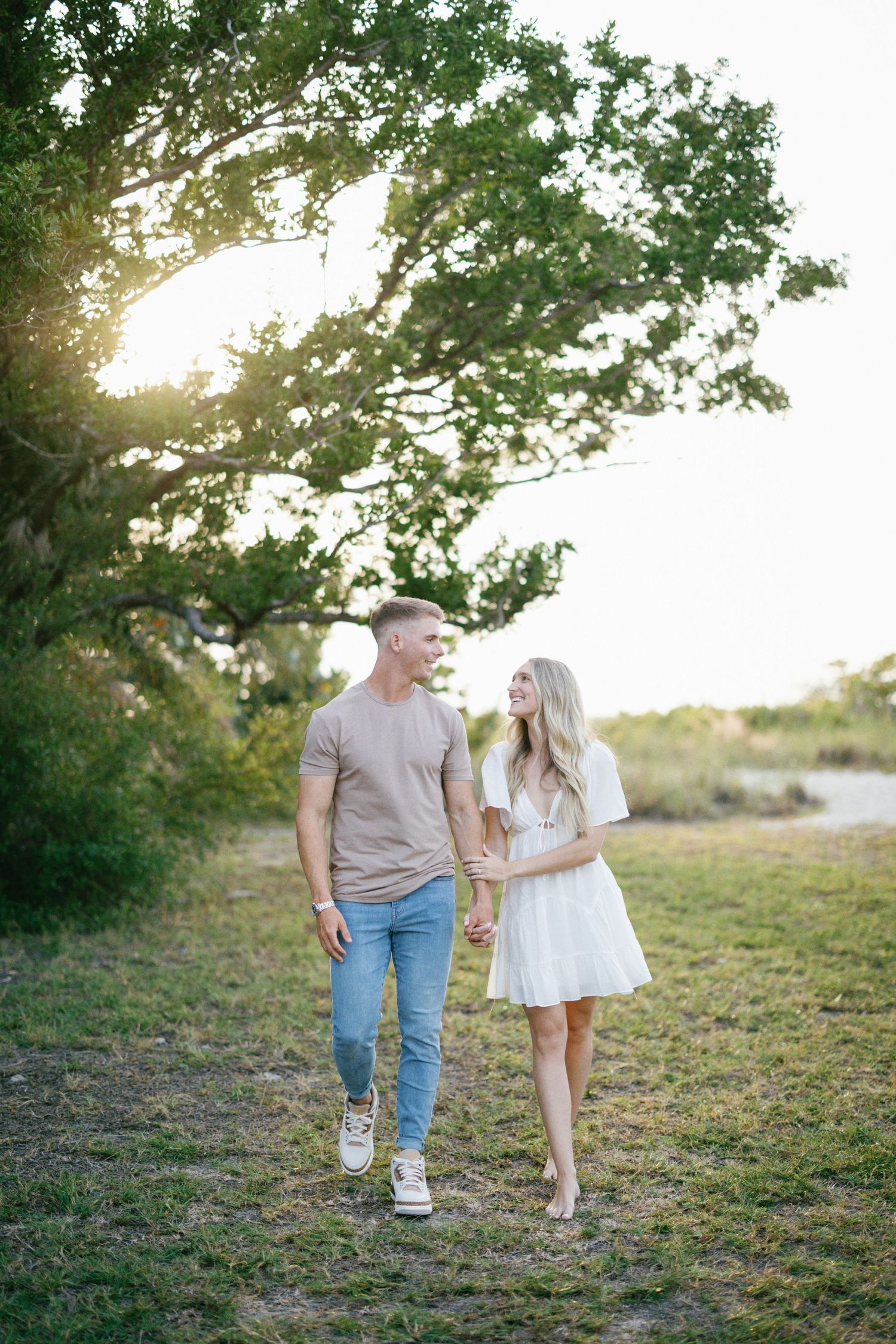 A couple takes a romantic stroll together through a grassy field at sunset during a photography session.