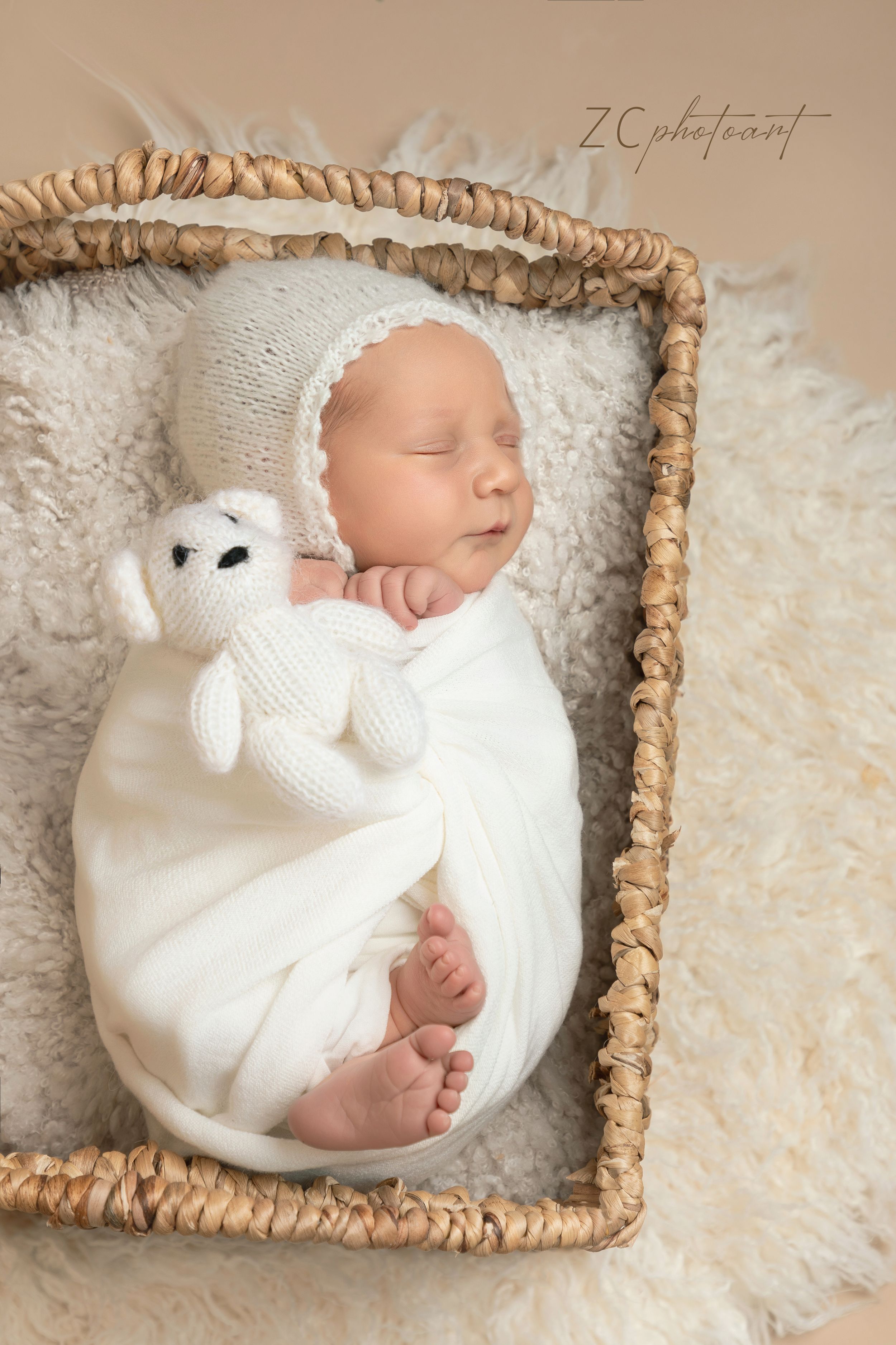 Sleeping newborn in white wrap lying on soft textured blanket in woven basket with delicate lighting and neutral tones.