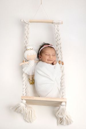 Newborn baby lying on decorative macrame swing with white rope tassels and wooden beams on light backdrop.
