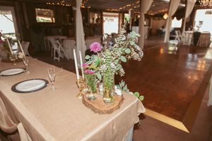 Rustic reception hall with wooden tables, white linens, and floral centerpieces in a barn-style setting.
