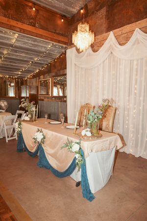 Rustic barn wedding reception setup with white drapes, chandeliers, and teal accents decorating the tables.