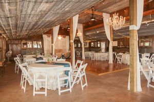 Decorated rustic event space with round tables, white chairs, and elegant chandeliers hanging from wooden ceiling.