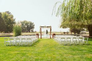 Outdoor wedding ceremony setup with white chairs and rustic wooden arch under drooping willow trees on green lawn.