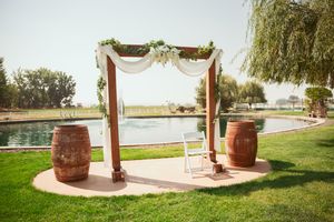 Outdoor wedding ceremony arch with white fabric and wine barrels on circular patio overlooking waterfront landscape.