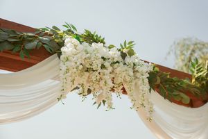 Elegant white floral and greenery arrangement draped across wooden beams with flowing fabric for wedding decor.