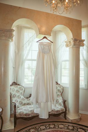 White christening gowns hanging in an elegant room with arched windows, ornate chandelier, and floral accent chair.