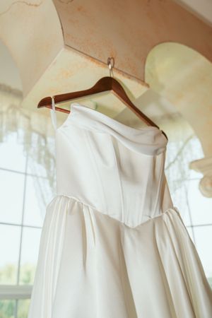 A row of elegant white christening gowns displayed on hangers against a sunny window backdrop.
