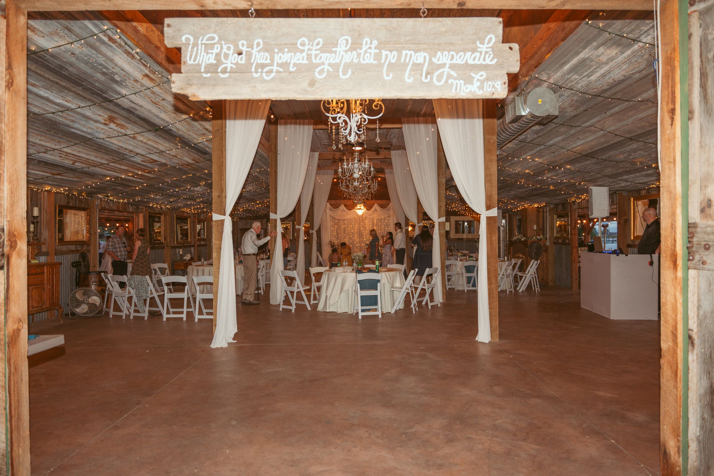 Rustic barn wedding venue with wooden beams, draped white curtains, chandeliers and white folding chairs set up for ceremony.