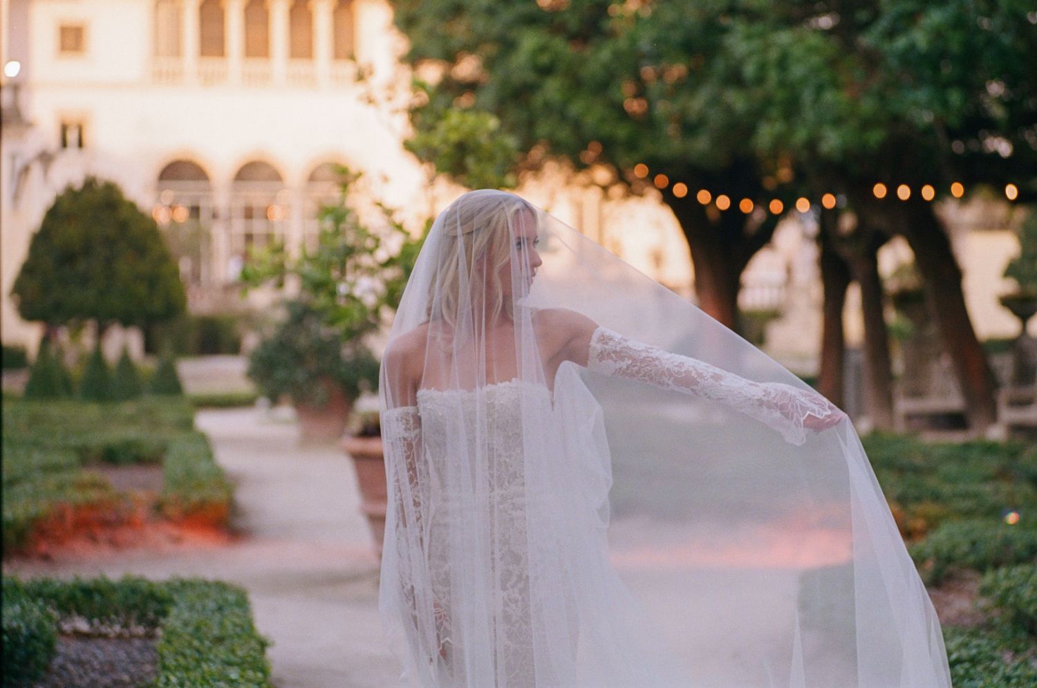 Elegant bride in a form-fitting lace wedding gown with sheer sleeves and flowing cathedral veil poses outdoors.