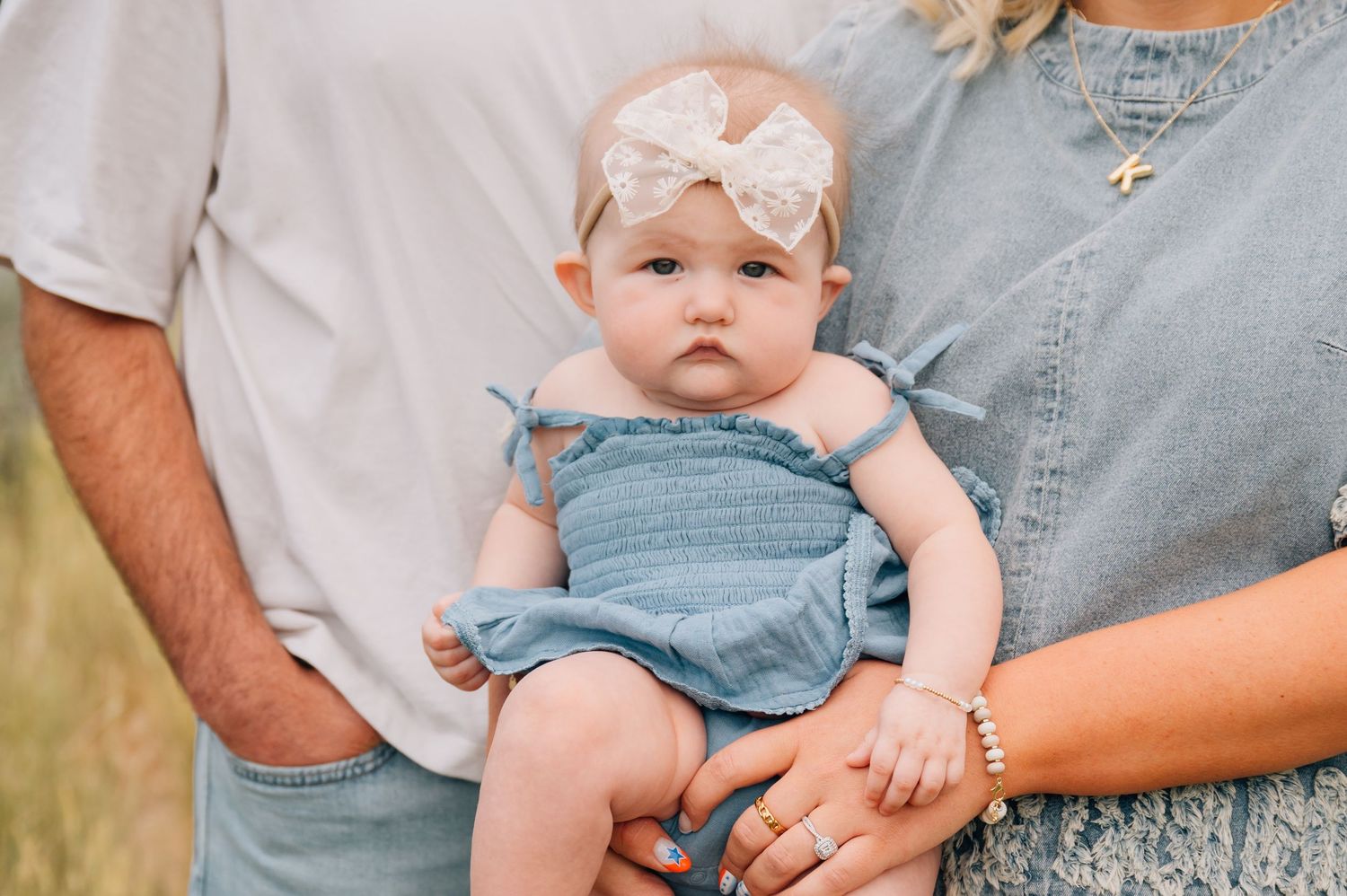 Infant wearing blue romper and white bow headband being held by parents in casual clothing outdoors.