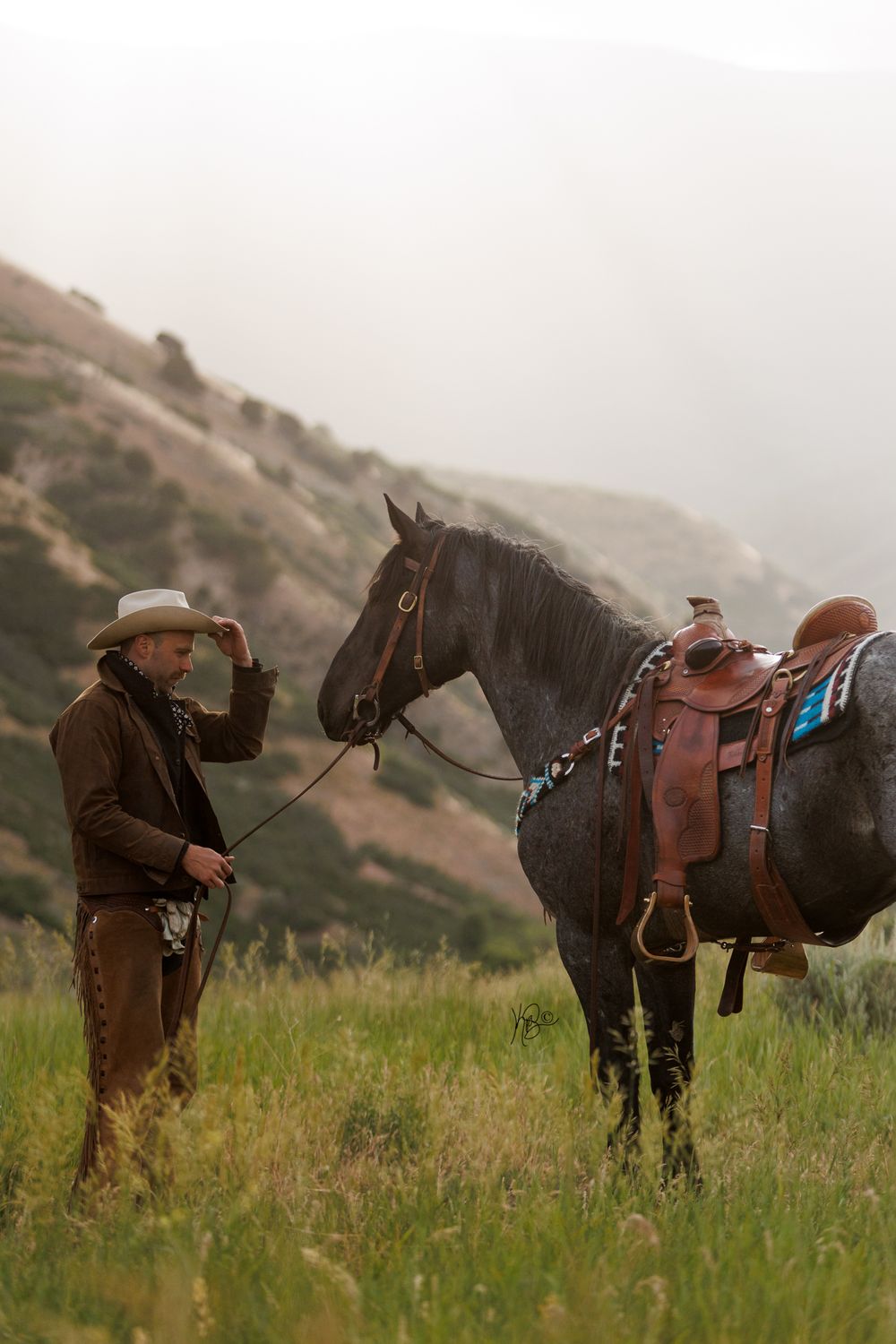 A cowboy stands beside a black horse with western tack against a mountainous backdrop.