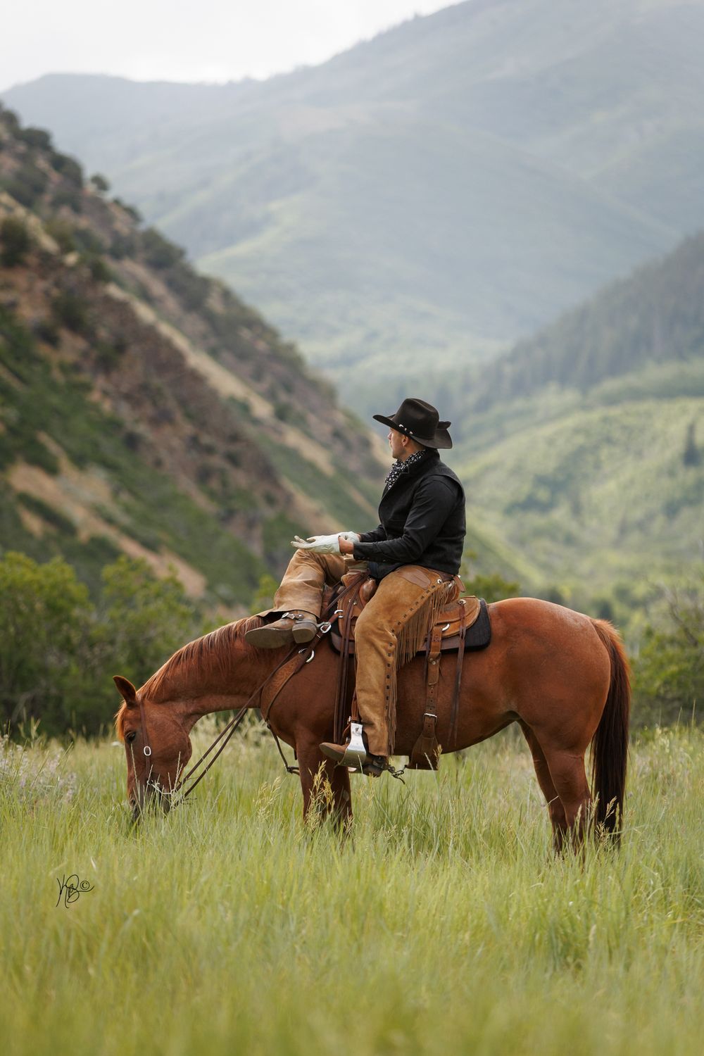 A horse grazes in tall grass while its rider takes in the mountain valley views.