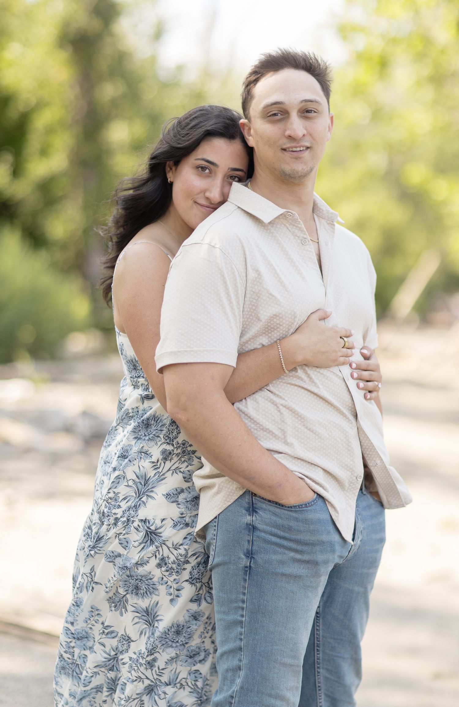 A couple embraces outdoors in natural lighting, with one wearing a floral dress and the other in a white shirt and jeans.