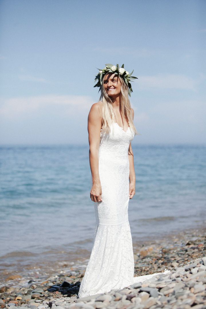 Woman in white lace dress walks along rocky beach shoreline on sunny day as waves lap at pebbled coast.